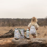 A child stands in a dry outdoor landscape wearing a matching backpack, with a patterned lunch bag and water bottle placed on a large log in the foreground.