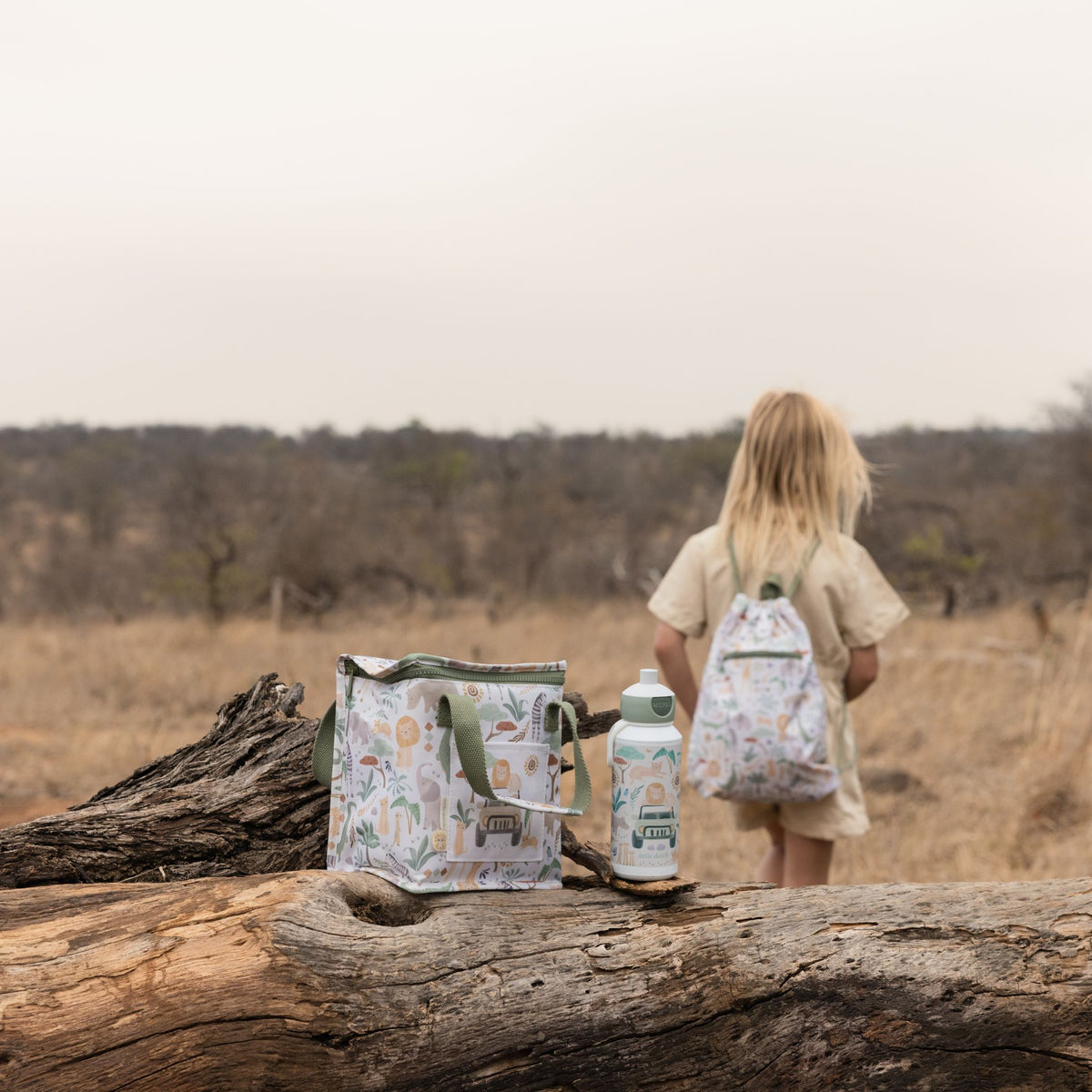A child stands in a dry outdoor landscape wearing a matching backpack, with a patterned lunch bag and water bottle placed on a large log in the foreground.