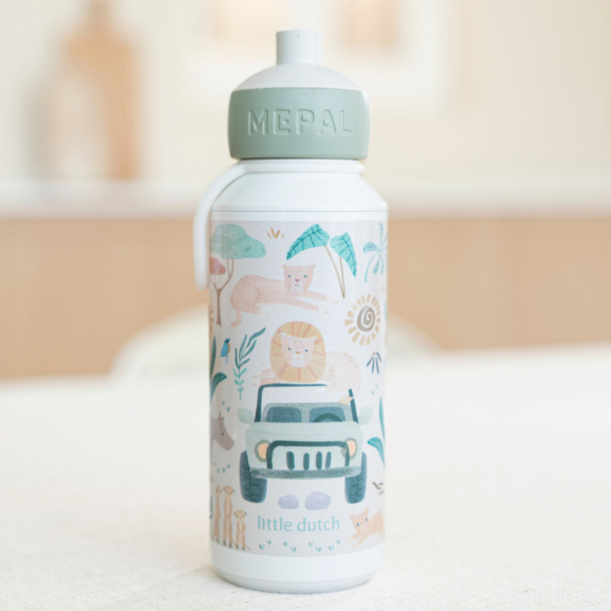 Little Dutch white Mepal water bottle with a light sage green lid, decorated with a whimsical safari illustration featuring a jeep, lion, elephant, and palm trees. The bottle is set against a softly blurred, warm-toned indoor background.
