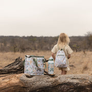 A child stands in a dry grassy outdoor landscape wearing a little dutch safari friends backpack while a matching insulated tote bag and water bottle sit on a fallen log in the foreground. The items have a light off-white base with a multicolor safari-style animal print.