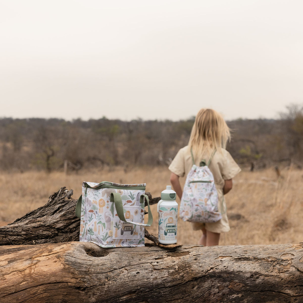A child stands in a dry grassy outdoor landscape wearing a little dutch safari friends backpack while a matching insulated tote bag and water bottle sit on a fallen log in the foreground. The items have a light off-white base with a multicolor safari-style animal print.