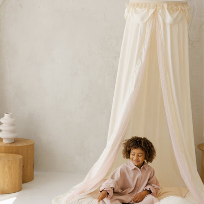Child sitting under a white canopy in a minimalistic room with a white wall and light-colored floor.