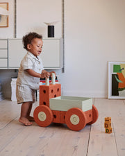 A toddler stands barefoot on a light wood floor with the MODU 3 in 1 Walker in colour burnt orange wheels. The child is wearing a light-colored short-sleeve shirt and shorts.