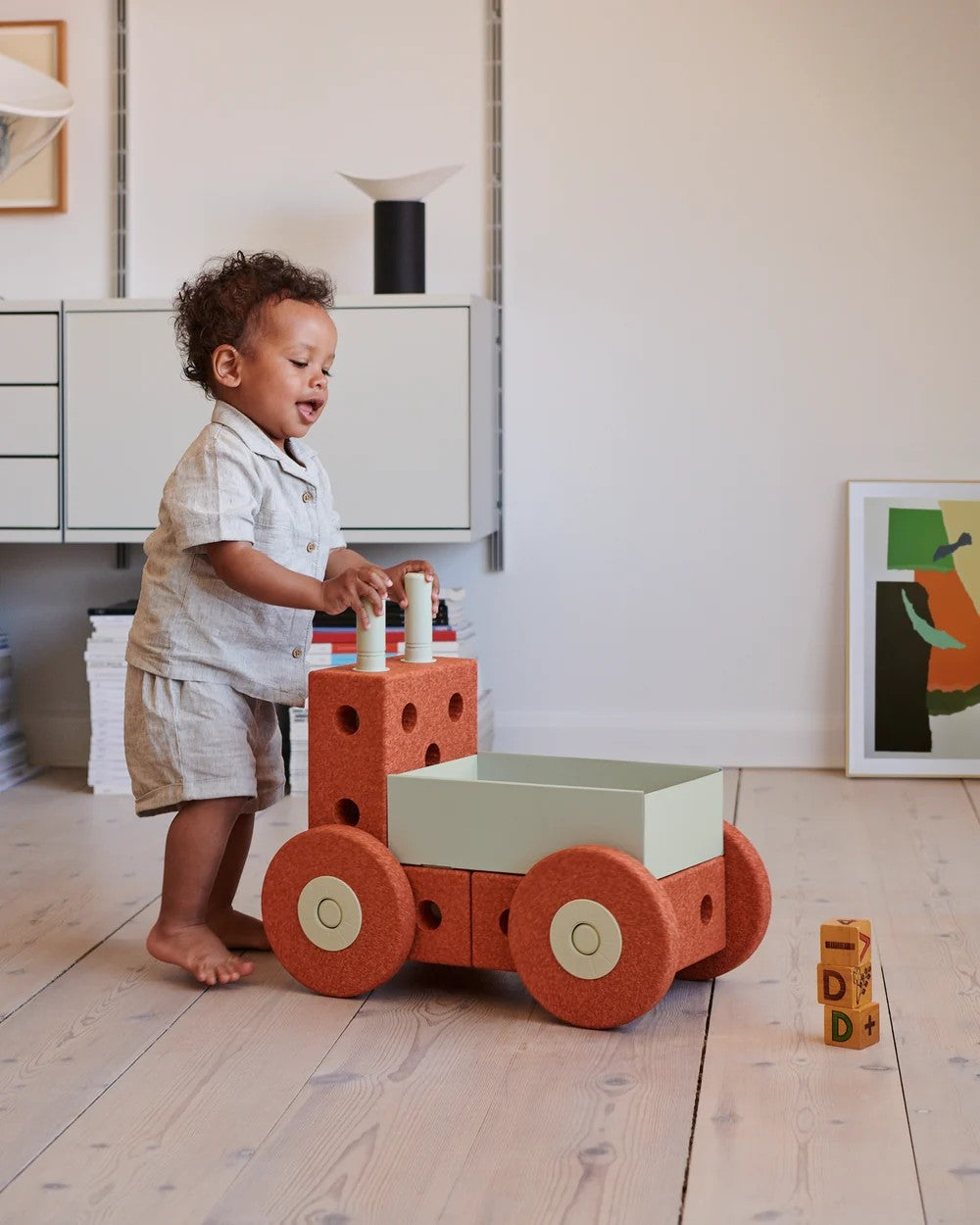 A toddler stands barefoot on a light wood floor with the MODU 3 in 1 Walker in colour burnt orange wheels. The child is wearing a light-colored short-sleeve shirt and shorts.