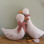 Two white fabric goose decorations with orange beaks and feet, wearing red patterned bows, displayed on a woven wicker surface against a pale green wall.