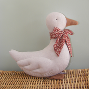 A plush fabric duck decoration in a pale blush pink color, featuring a brown fabric beak and feet, and a red and white floral patterned neck bow, displayed on a woven wicker surface against a soft sage green background.