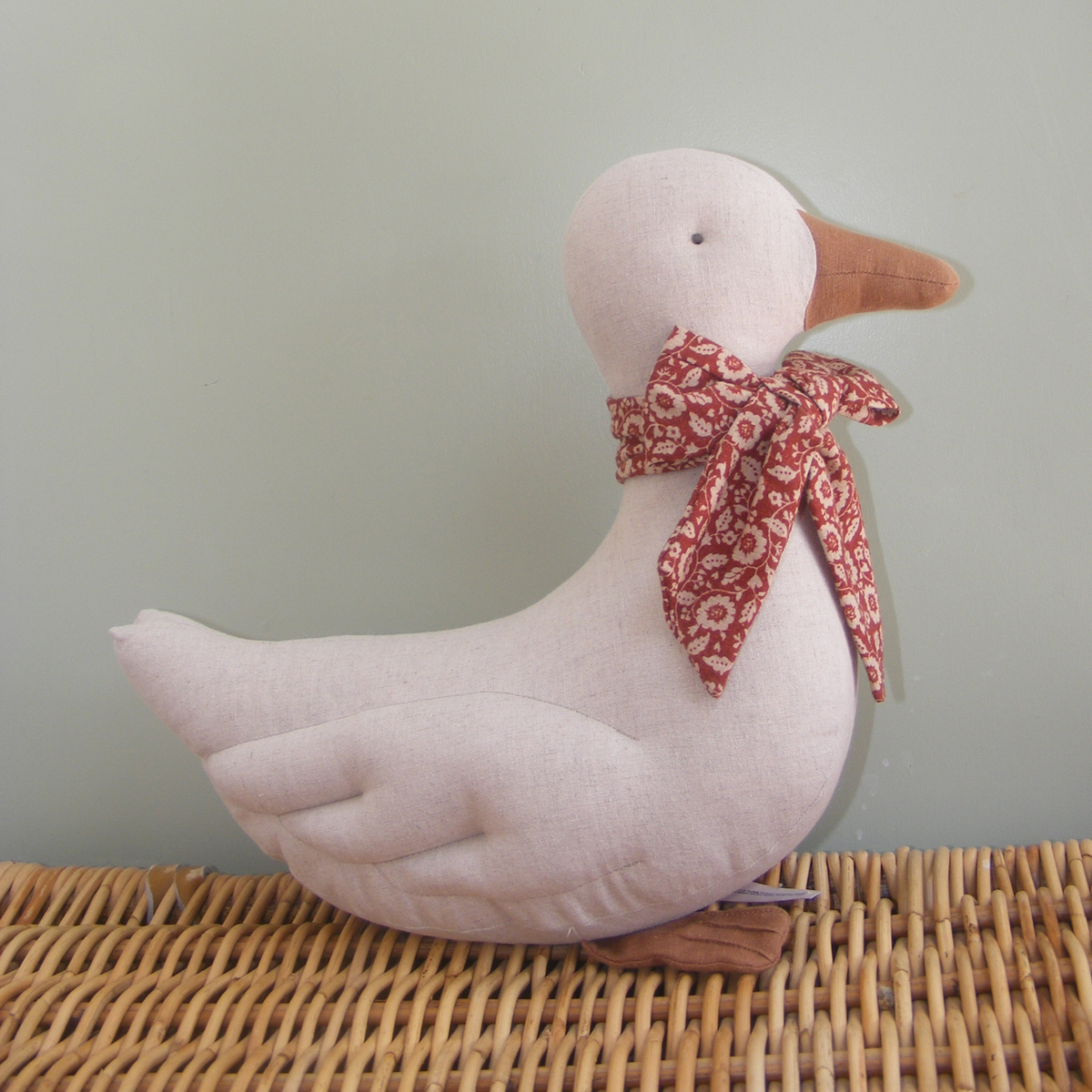 A plush fabric duck decoration in a pale blush pink color, featuring a brown fabric beak and feet, and a red and white floral patterned neck bow, displayed on a woven wicker surface against a soft sage green background.