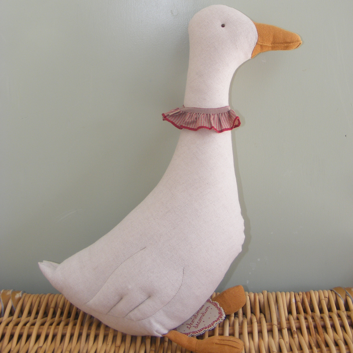 A handmade fabric goose plush toy with a white body, orange beak and feet, and a pink ruffled collar, sitting on a woven wicker surface against a muted green wall.