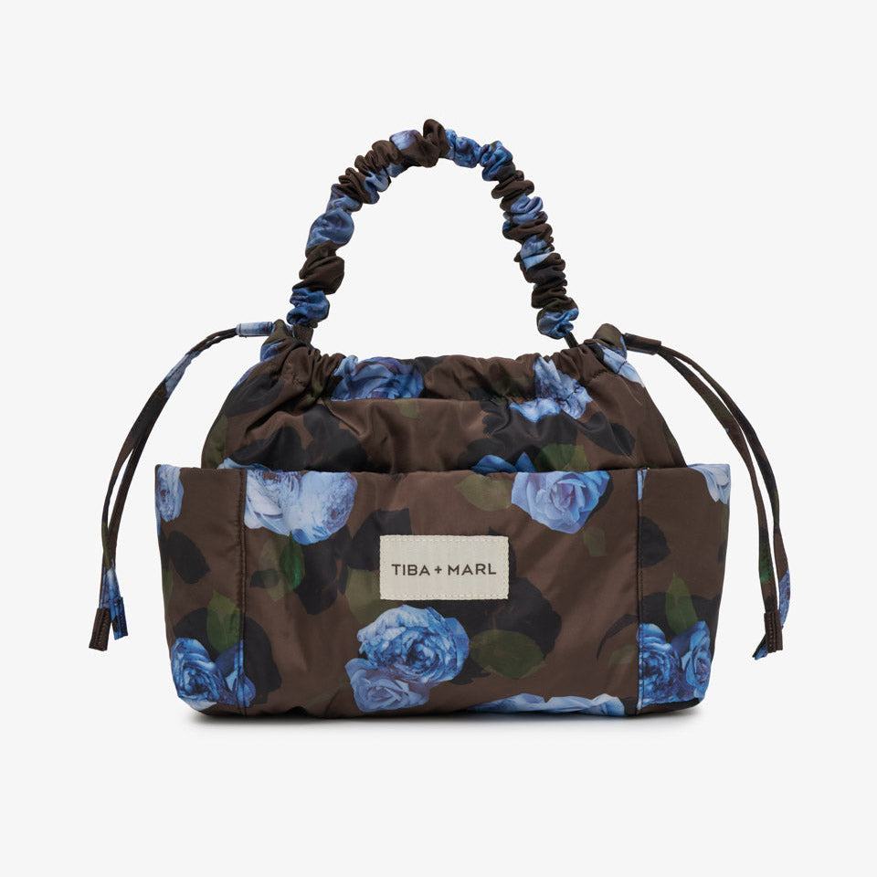 A Tiba + Marl handbag with a brown base and blue floral print, featuring a gathered top handle and a front patch logo.