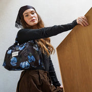 A woman wearing a black headscarf, black long-sleeve top, and brown cargo pants carries a black nylon duffel bag with a blue floral print. She is leaning against a wooden panel in a minimalist room.