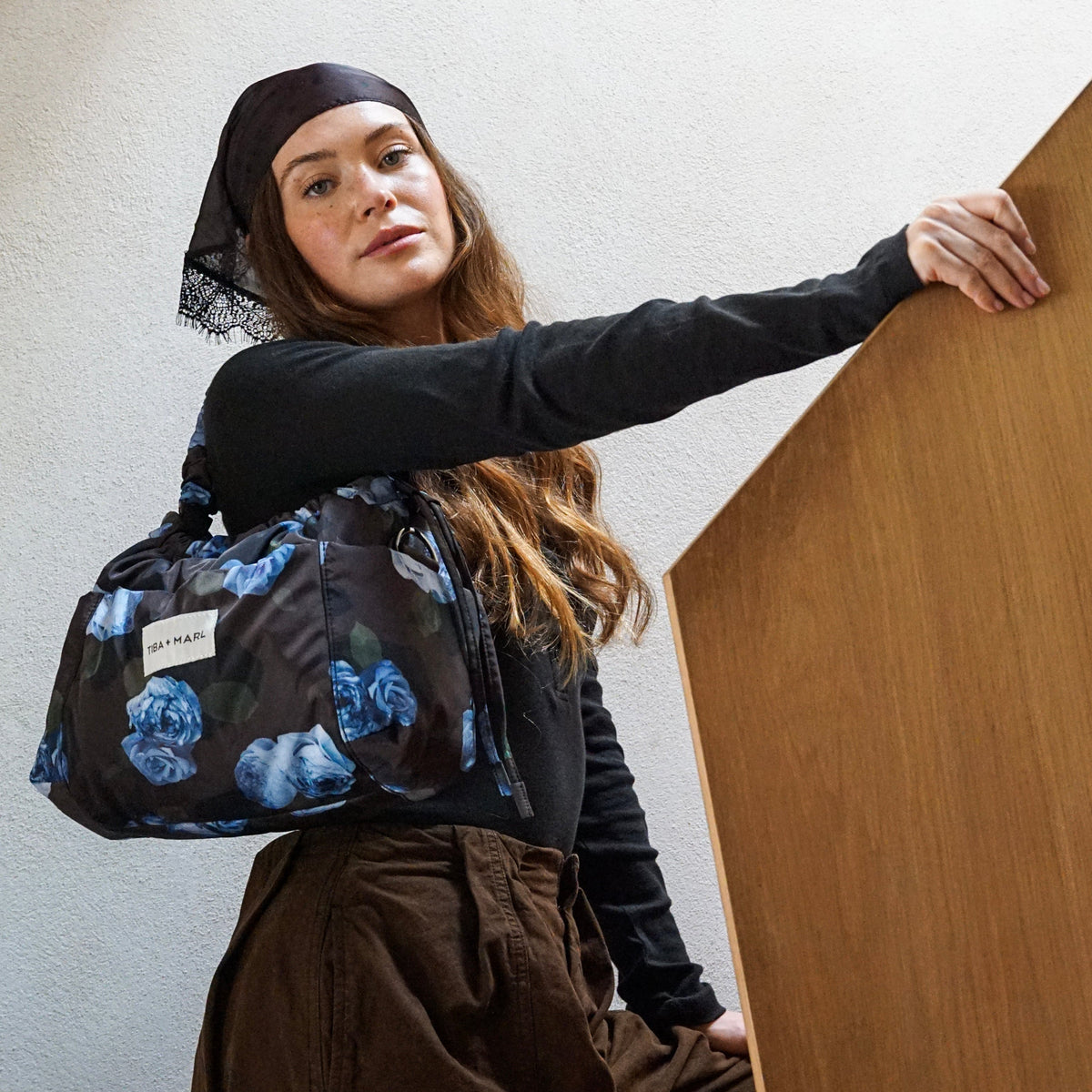 A woman wearing a black headscarf, black long-sleeve top, and brown cargo pants carries a black nylon duffel bag with a blue floral print. She is leaning against a wooden panel in a minimalist room.