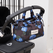 A floral drawstring pouch bag by Tuba & Karl, featuring blue roses on a dark background, hanging from a black stroller handle.