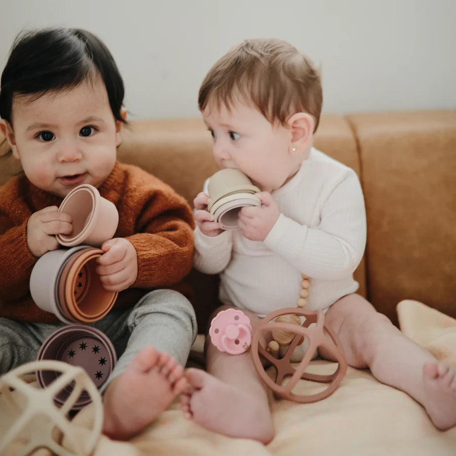 Two babies playing with mushie toys on a sofa