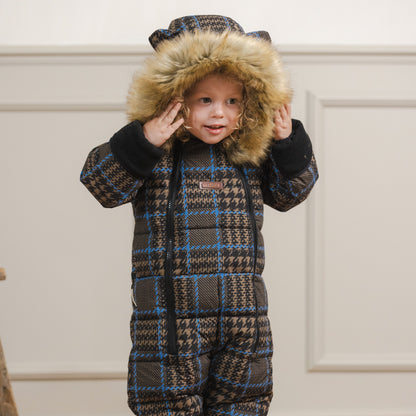 A child wearing a full-body winter snowsuit with a brown, black, and blue plaid pattern, a faux-fur trimmed hood, and tan boots, standing in an indoor room with white wall paneling and light wood flooring.