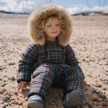 A young child wearing a brown and blue plaid hooded winter suit with a faux-fur trimmed hood sits on a sandy, pebble-strewn beach under a cloudy blue sky.