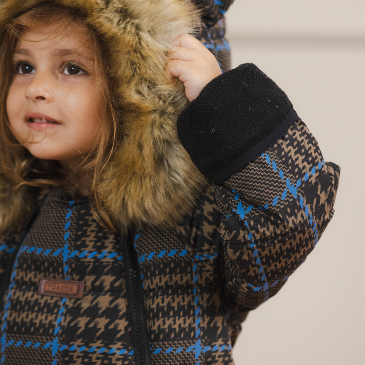 A child wearing a brown and black houndstooth patterned winter snowsuit with bright blue grid lines, a black ribbed collar, and a brown faux-fur trimmed hood. The child is holding up the hood.