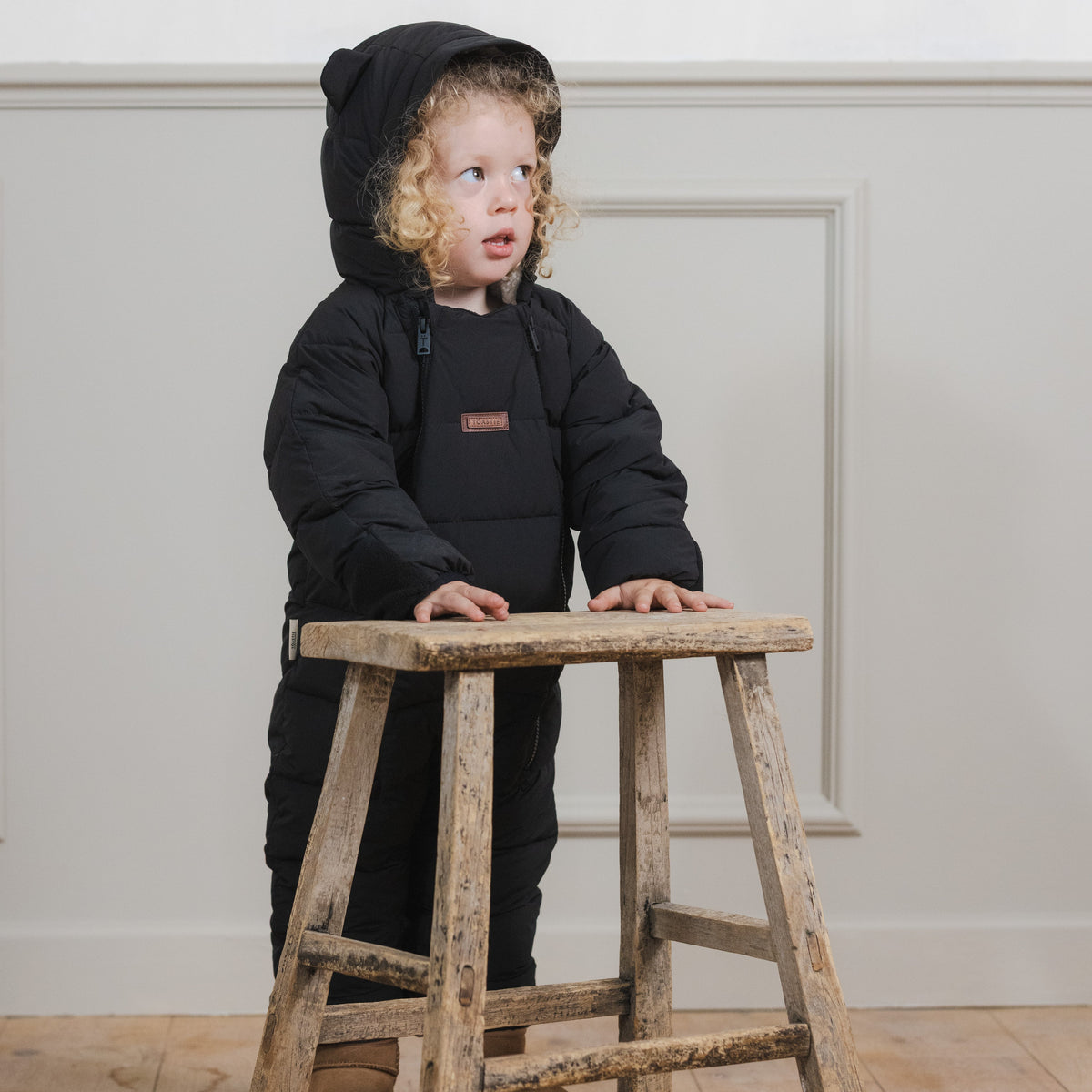 A toddler wearing a black hooded puffer snowsuit stands behind a rustic wooden stool in a bright room with a light wall and wood floor.