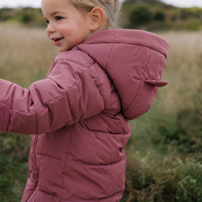 A young girl with blonde hair in a ponytail wearing a dusty rose pink hooded puffer snowsuit, standing in an outdoor grassy field under an overcast sky.