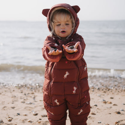 A toddler stands on a sandy beach wearing a dark reddish-brown hooded puffer snowsuit with small white horse prints. The background shows the ocean and a pale sky.