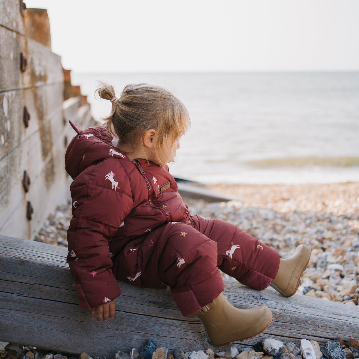 A toddler sits on a weathered wooden board on a pebble beach, wearing a deep burgundy puffer snowsuit with a white horse print, paired with tan boots.