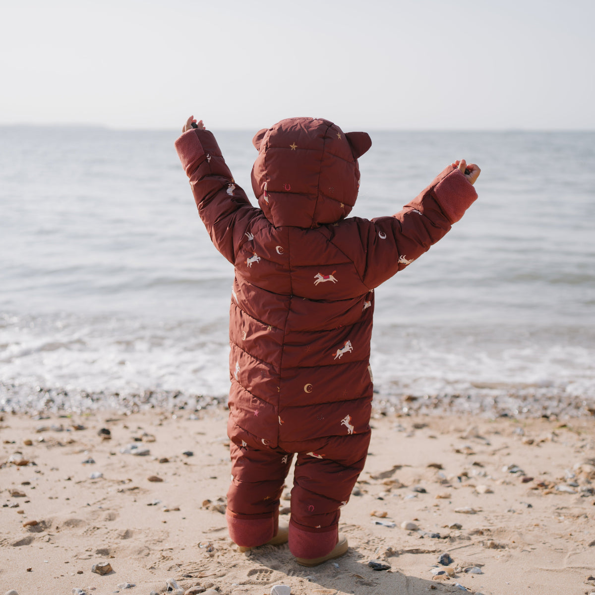 A baby wearing a rust-red hooded snowsuit stands on a sandy beach with the ocean in the background, facing the water with arms raised.