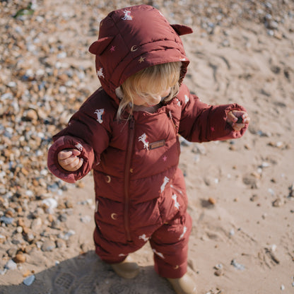 A toddler standing on a sandy beach wearing a dark reddish-brown puffer snowsuit with white unicorn and moon prints, featuring a hood with bear ears.