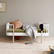 Modern Scandinavian Oliver Furniture Wood Original Junior Bed in white and oak with curved corners, featuring a green mattress, pink pillow, white bedding, and wooden floor in a minimalist nursery room.