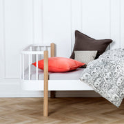 Modern white and oak Scandinavian-style Oliver Furniture Wood Original Bed with curved corners, featuring elegant 1950s design, positioned next to a parent bed in a minimalist bedroom with patterned bedding and decorative pillows.