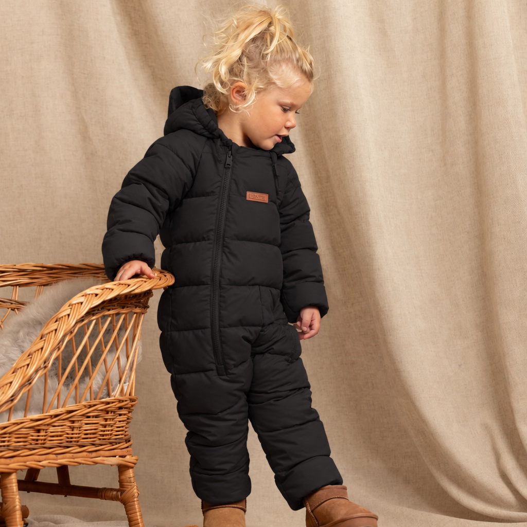 A toddler stands in a studio setting wearing a black puffer snowsuit and brown boots, leaning against a wicker chair in front of a beige draped backdrop.