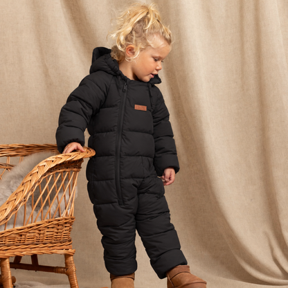 A toddler stands in a studio setting wearing a black puffer snowsuit and brown boots, leaning against a wicker chair in front of a beige draped backdrop.
