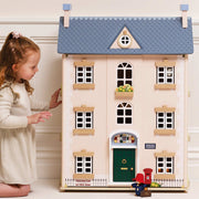 A young girl in a cream dress stands beside a large wooden dollhouse-style house with a blue roof, cream facade, green door, and small street details, photographed indoors against a light wall.