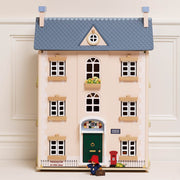 A wooden le toy van dolls house featuring a classic three-story pink building with a blue patterned roof, green front door, red postman figure, and red mailbox, set against a cream paneled wall.