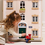 A young girl with curly brown hair kneels beside a large le toy van wooden dolls house, reaching toward a green front door. The dolls house has cream-colored siding, white-framed windows, a small flower box, and a blue sign reading 'WINDSOR GARDENS'.