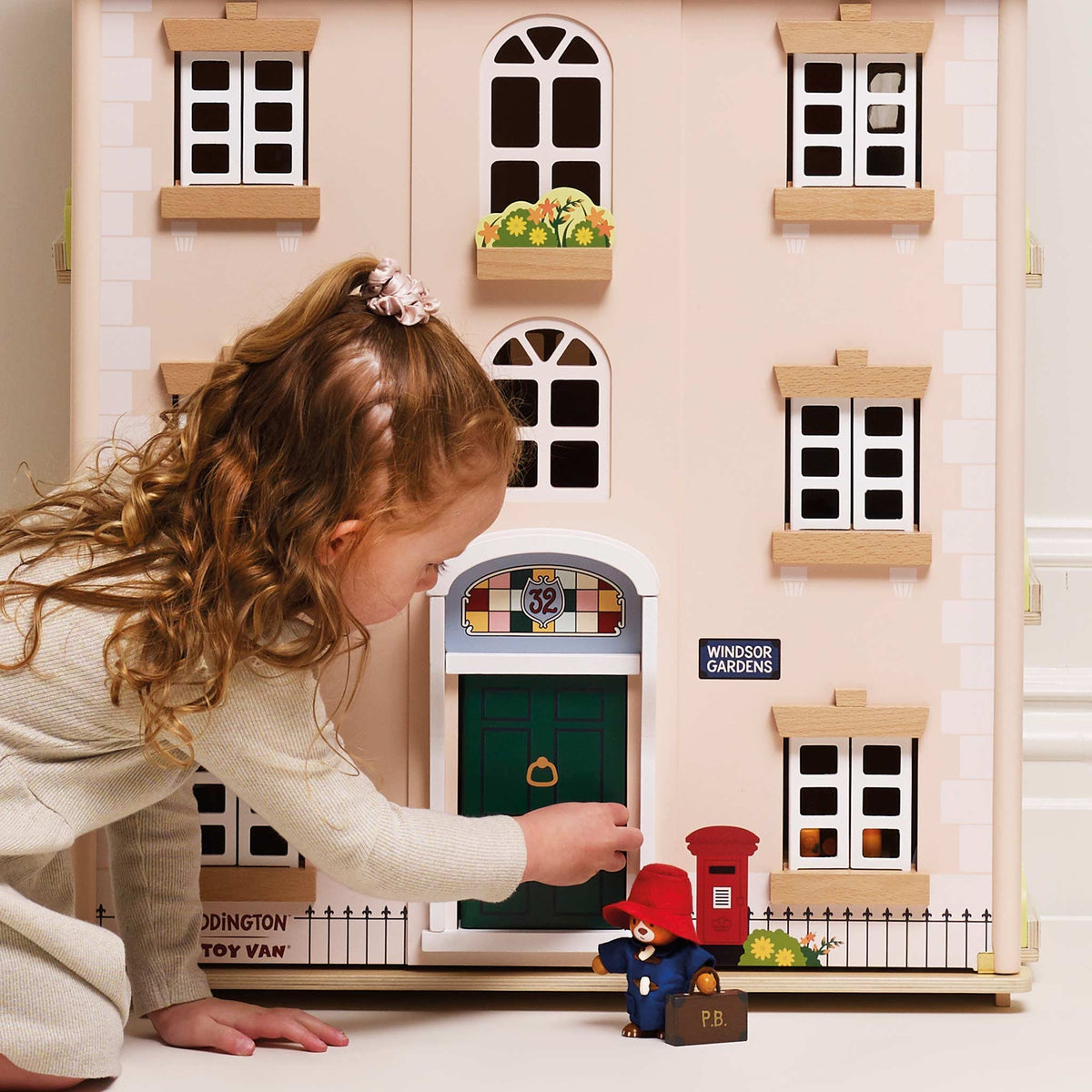 A young girl with curly brown hair kneels beside a large le toy van wooden dolls house, reaching toward a green front door. The dolls house has cream-colored siding, white-framed windows, a small flower box, and a blue sign reading 'WINDSOR GARDENS'.