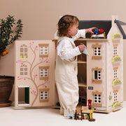 A toddler in cream overalls plays with a large wooden dollhouse with an open front door showing a tree illustration, set against a warm beige background.
