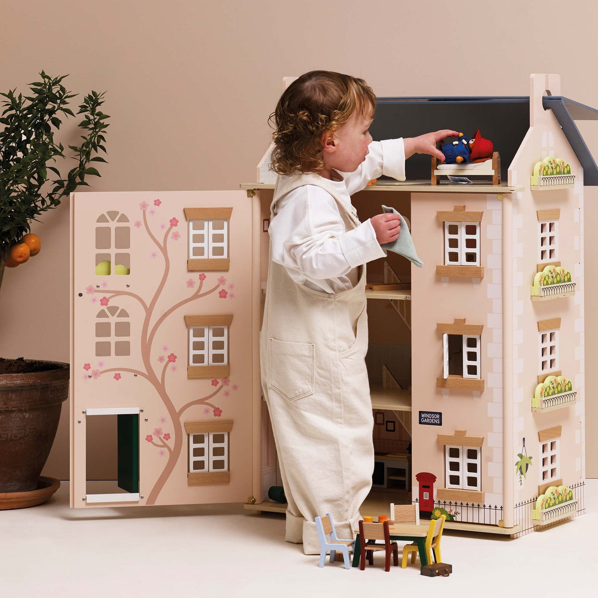 A toddler in cream overalls plays with a large wooden dollhouse with an open front door showing a tree illustration, set against a warm beige background.