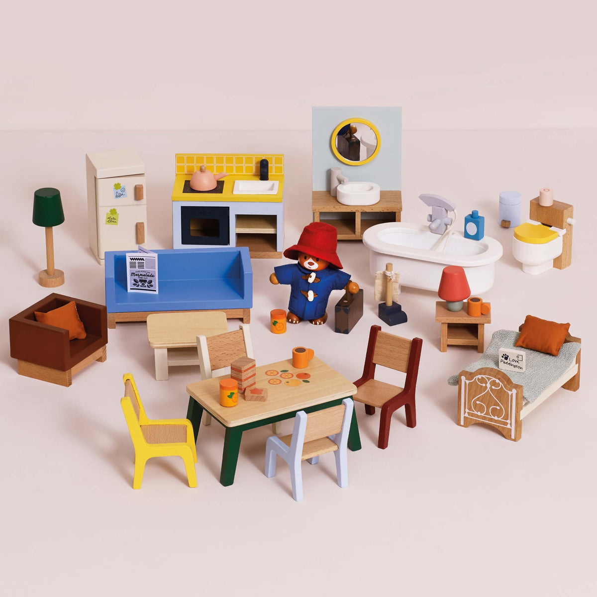 A miniature wooden dollhouse furniture set arranged on a pale pink background. The set includes a kitchen, bathroom, living room, bedroom, and dining area with small wooden furniture pieces, chairs, tables, lamps, and a small figurine.