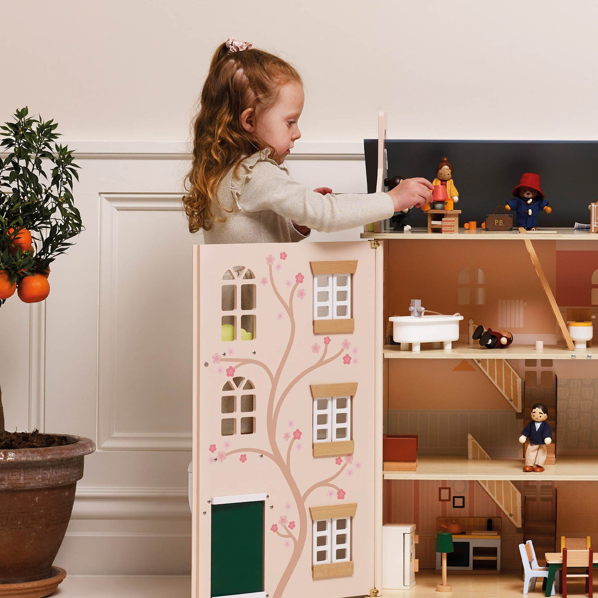A young girl with long wavy light brown hair stands behind a large wooden dollhouse, reaching toward a figure on the dollhouse roof. The dollhouse has a cream-colored exterior with pink cherry blossom tree illustrations, and an open interior showing multiple levels, stairs, and miniature furniture.