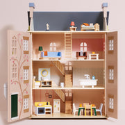 A miniature dollhouse with open doors, showing multiple interior floors with furniture, stairs, a kitchen, bathroom, and a small roof top, photographed against a plain light beige background.