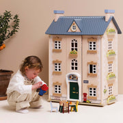 A child in a white outfit crouches beside a large dollhouse-style house with a blue roof and cream facade, playing with small furniture and toy accessories in a softly lit indoor setting.