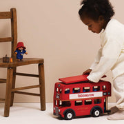 A young child in a cream outfit pushes a red double-decker toy bus labeled PADDINGTON. A wooden chair on the left holds a small Paddington figurine against a plain beige studio background.