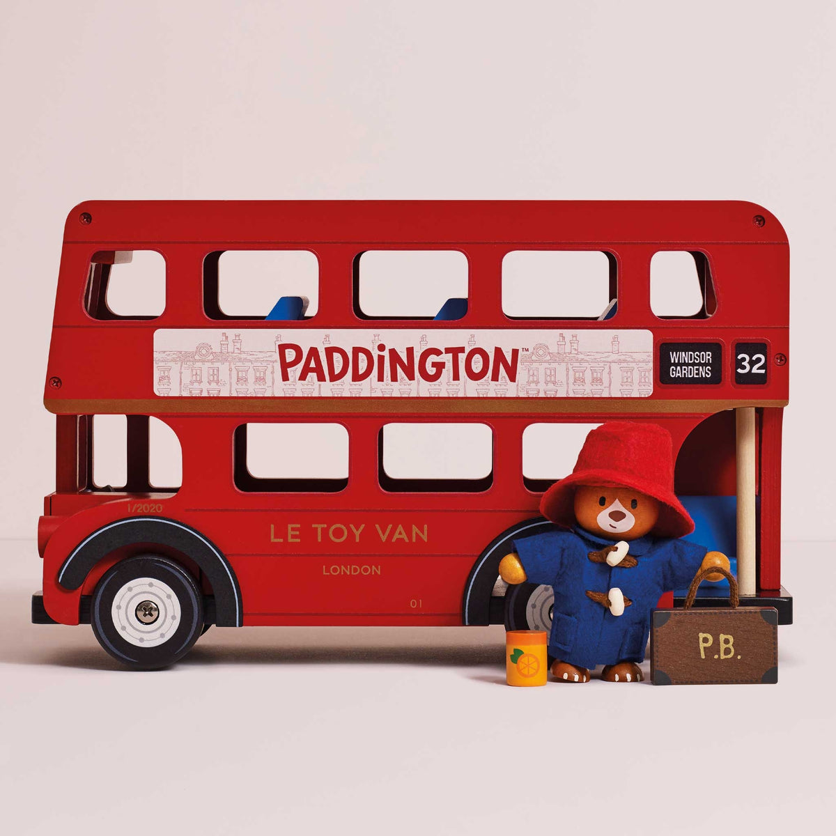 A red double-decker toy bus with Paddington Bear figurine and accessories, photographed on a clean light beige studio background.