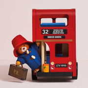 A red wooden double-decker bus toy with Paddington Bear figurine, dressed in a blue coat and red hat, standing beside it holding a suitcase. The bus has '32 Windsor Gardens' and 'LTV 1995' markings.