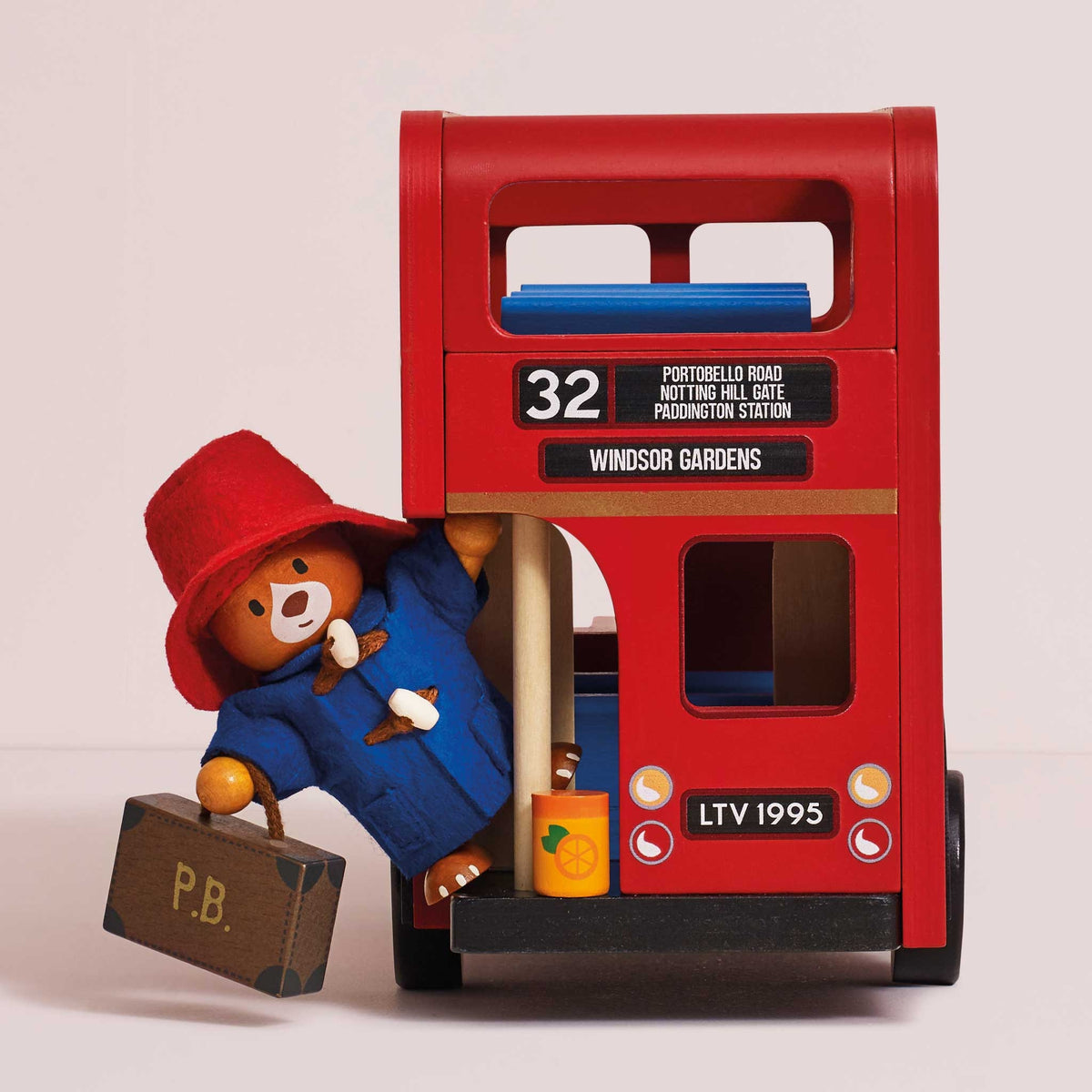 A red wooden double-decker bus toy with Paddington Bear figurine, dressed in a blue coat and red hat, standing beside it holding a suitcase. The bus has '32 Windsor Gardens' and 'LTV 1995' markings.
