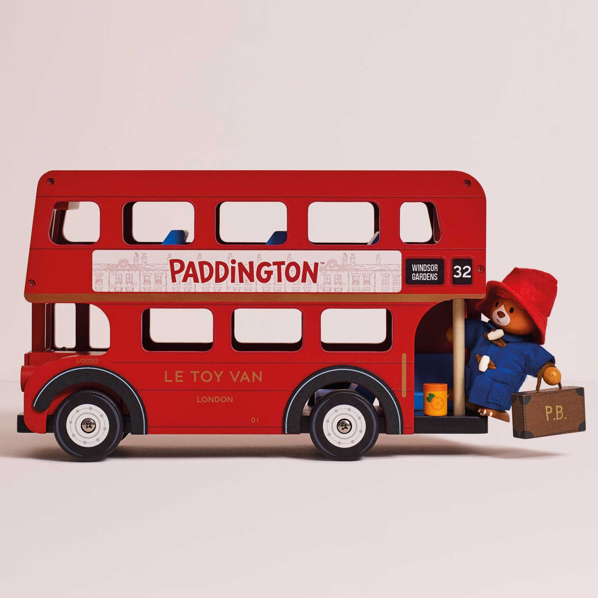 A red wooden double-decker toy bus with Paddington Bear seated at the open rear door, holding a brown suitcase. The bus has 'PADDINGTON' and 'LE TOY VAN LONDON' markings, with Paddington wearing a red hat and blue coat.