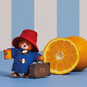 A Paddington Bear figurine wearing a red hat and blue coat, holding an orange and standing beside a small brown suitcase, with a real orange half and a whole orange on a blue surface against a blue-and-white striped background.