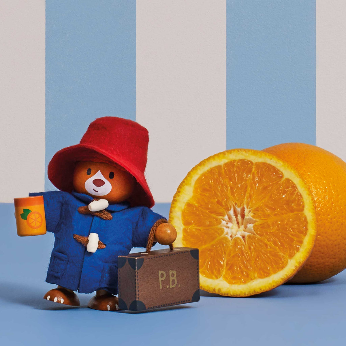 A Paddington Bear figurine wearing a red hat and blue coat, holding an orange and standing beside a small brown suitcase, with a real orange half and a whole orange on a blue surface against a blue-and-white striped background.