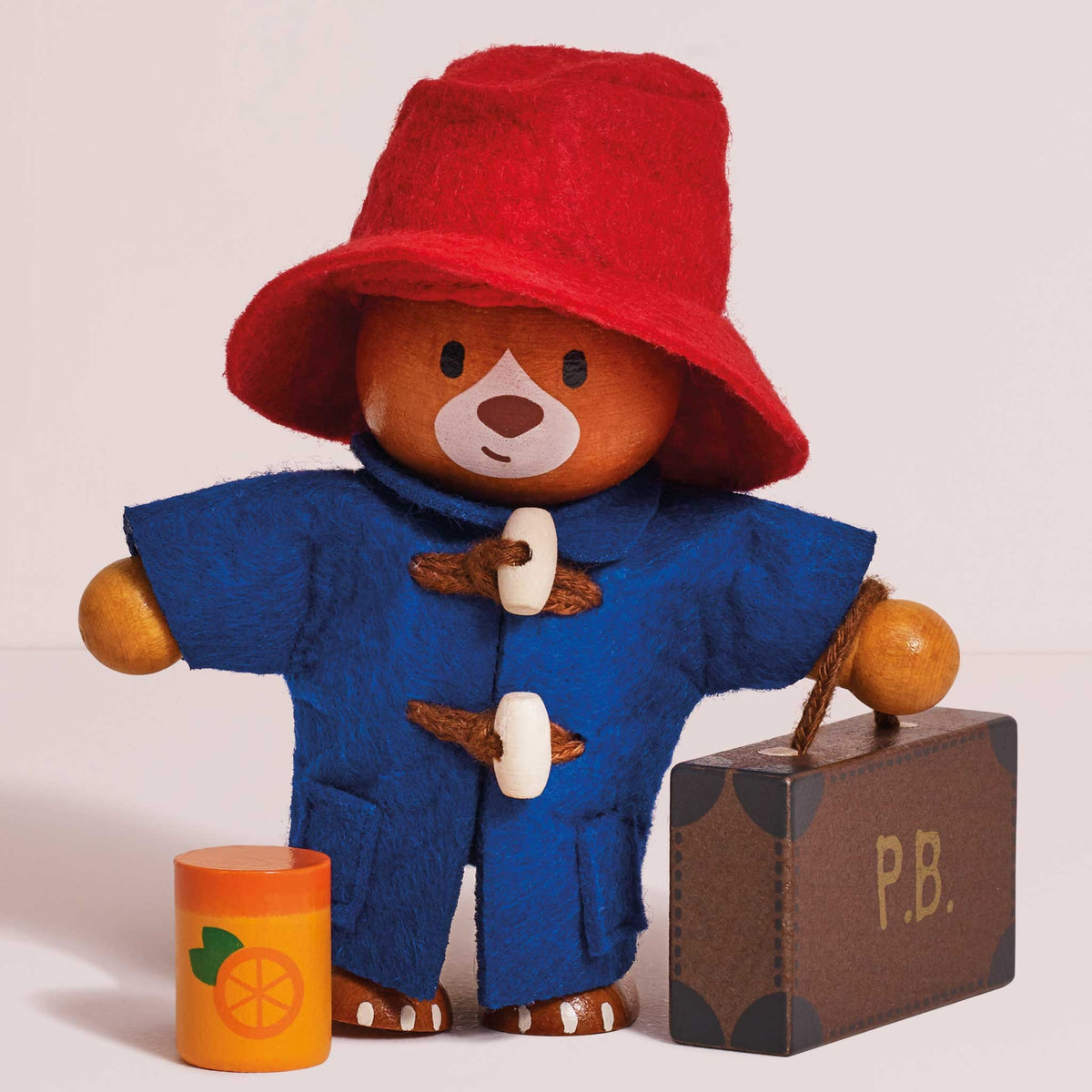 A wooden Paddington Bear toy wearing a red felt hat and blue felt duffle coat, standing beside a brown suitcase marked P.B.