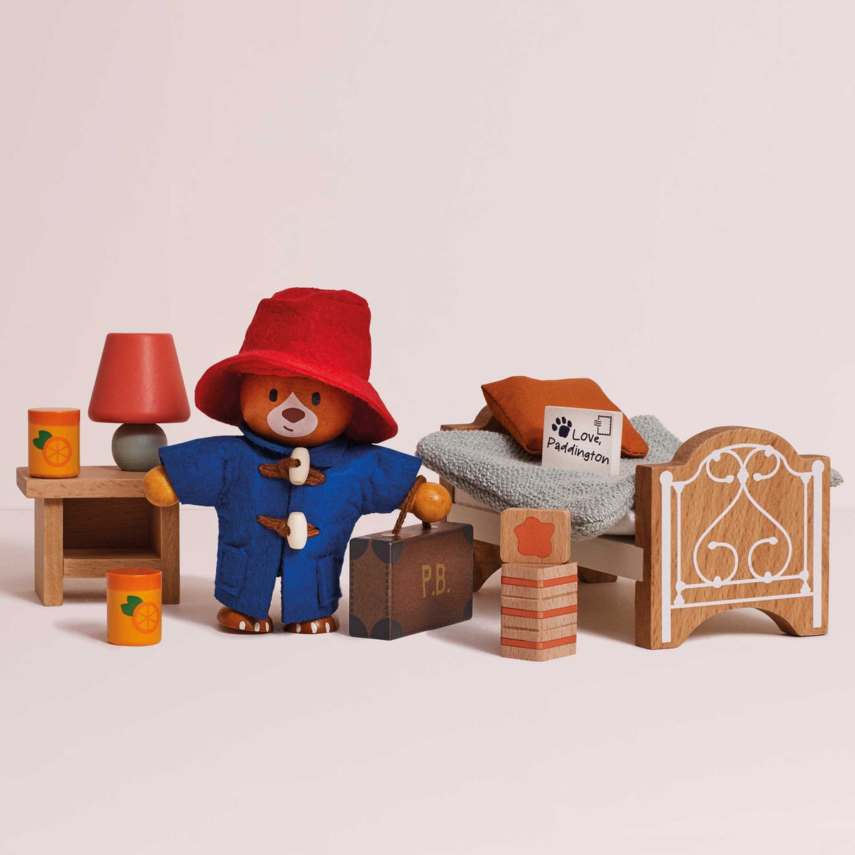 A wooden Paddington Bear toy set arranged on a light beige background, including a red hat, blue coat, suitcase, bedside table with lamp, bed with gray bedding and orange pillow, and small wooden blocks.