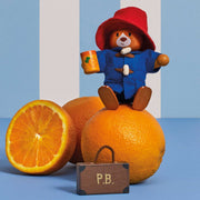 A Paddington Bear wooden doll wearing its signature red hat and blue duffel coat, sitting on top of a whole orange and holding an orange mug. A cut orange half and a small brown suitcase labeled 'P.B'
